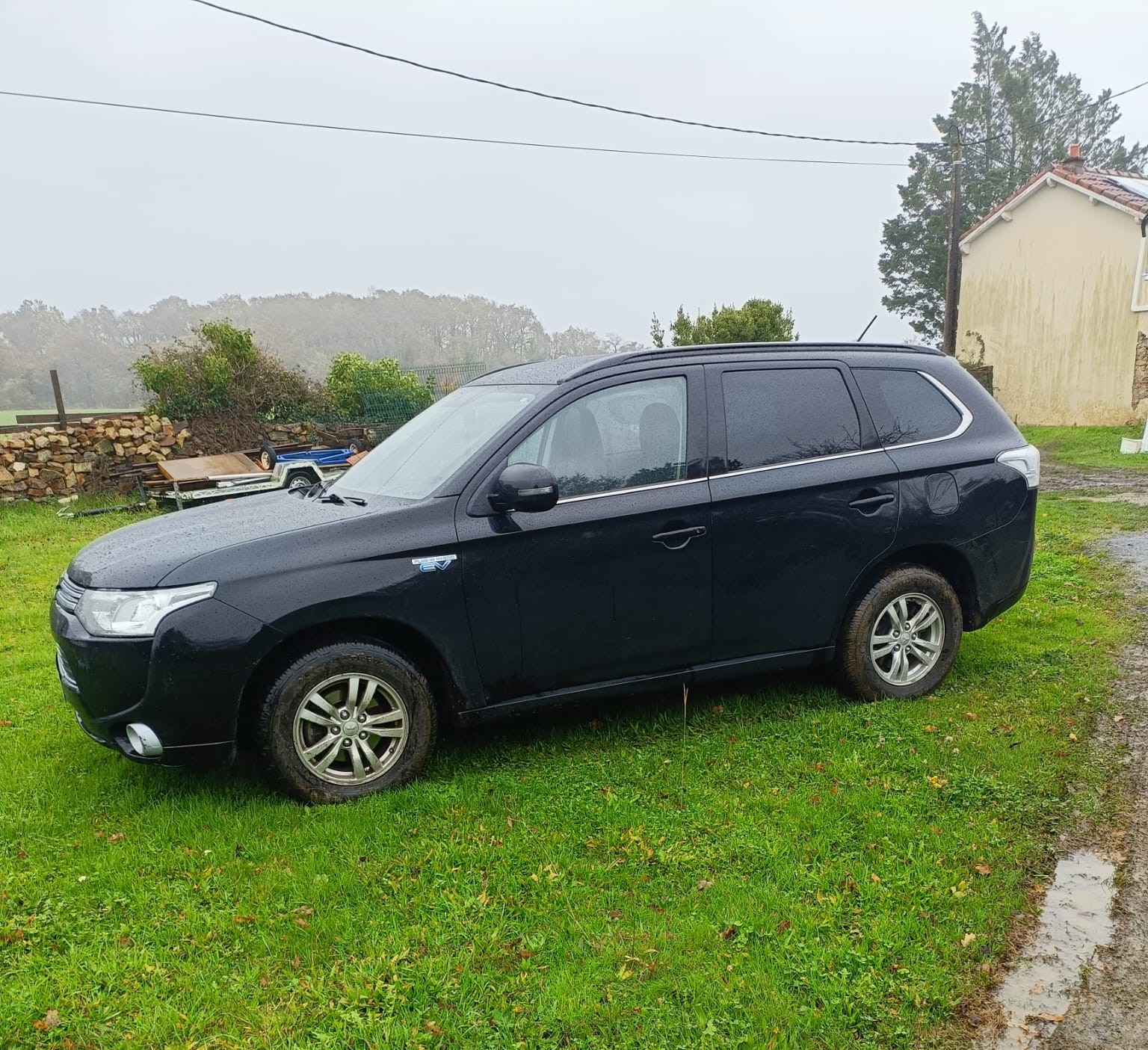 Mitsubishi Outlander PHEV noir de profil côté conducteur, stationné sur l’herbe avec une maison et des arbres en arrière-plan