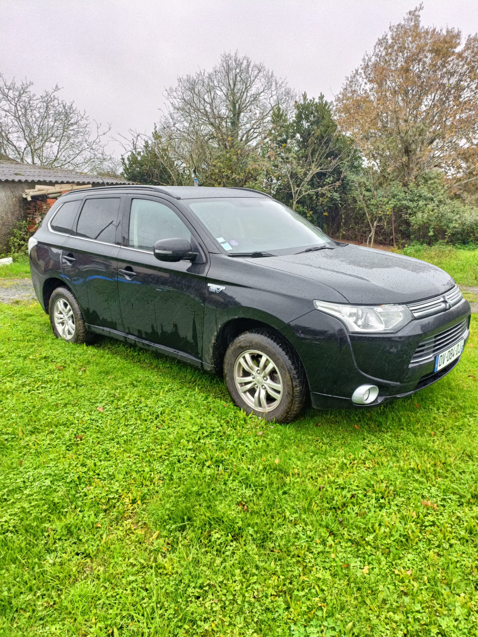 Mitsubishi Outlander PHEV noir vu de trois-quarts avant gauche, roues légèrement sales, sur une pelouse verte en milieu rural